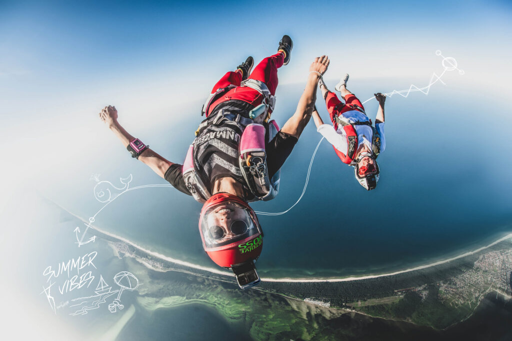 Dwóch spadochroniarzy w kolorowych kombinezonach i kaskach spada swobodnie nad wybrzeżem; na zdjęciu widoczne są białe rysunki oraz napis „letni klimat”.